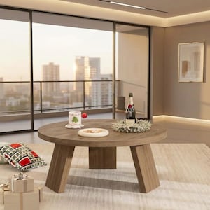 May include: A round wooden coffee table with three angled legs. On the table sits a white mug with a green tree graphic, a small white plate, a bottle of champagne, and a decorative wreath. A gift box and a patterned pillow are nearby.