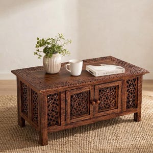 May include: A rectangular dark brown wooden coffee table featuring intricate carved patterns on the top surface and side panels. The table includes two central cabinet doors with small round knobs and a white ceramic vase with a green plant.