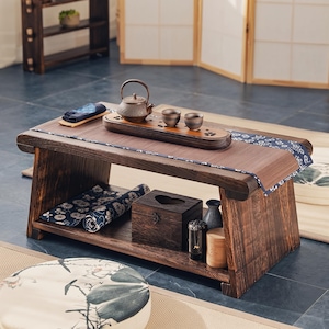 May include: A low rectangular wooden tea table with a dark brown finish, featuring a top surface with a blue and white patterned runner, a cast iron teapot, two small ceramic cups on a wooden tray, and a lower shelf holding a wooden box.