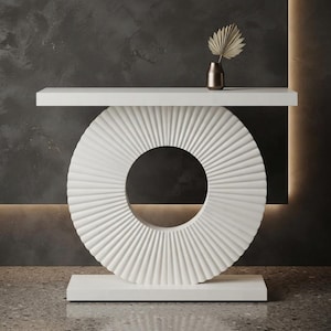 May include: A white console table featuring a flat rectangular top and a base composed of a large, circular, vertically fluted disc resting on a rectangular plinth. A small, dark metallic vase containing a single dried palm leaf sits on top.