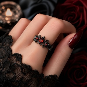 May include: A black metal ring featuring a crown-like design with intricate filigree detailing. The band is set with multiple small, faceted red stones, including a prominent heart-shaped red stone at the center of the crown motif.