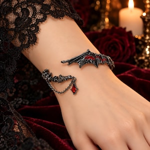 May include: A black metal cuff bracelet featuring a detailed bat wing design with red accents. The bracelet includes a textured bone-like structure on one side, connected by a thin black chain to a small, diamond-shaped red gemstone charm.