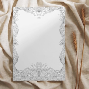May include: A rectangular, white paper with an ornate black and white floral border. The design features detailed patterns of leaves, swirls, and floral elements. The paper is set against a neutral-toned fabric background, with two stalks of wheat.