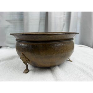 May include: A round, hammered brass bowl with a dark, aged patina. The bowl features a wide, flat rim and is supported by three ornate, claw-style feet with intricate detailing. The surface has a textured, dimpled appearance throughout.