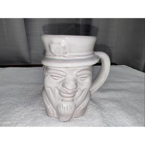 May include: A white ceramic figural mug shaped like a person's head wearing a hat with a small clover emblem on the front. The mug features a molded face with closed eyes, a smiling mouth, and a goatee, complete with a curved handle on the right side.