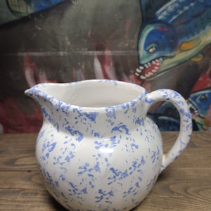 May include: A white ceramic pitcher with a blue and white speckled pattern. The pitcher has a rounded body and a curved handle.