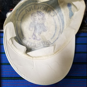 Op de afbeelding: Witte pet met een gebogen klep, met een blauwe afbeelding van een ram en de woorden "CAROLINA TAR HEELS". De binnenkant heeft een bedrukt ontwerp. De pet ligt op een gestreept blauw en groen oppervlak.