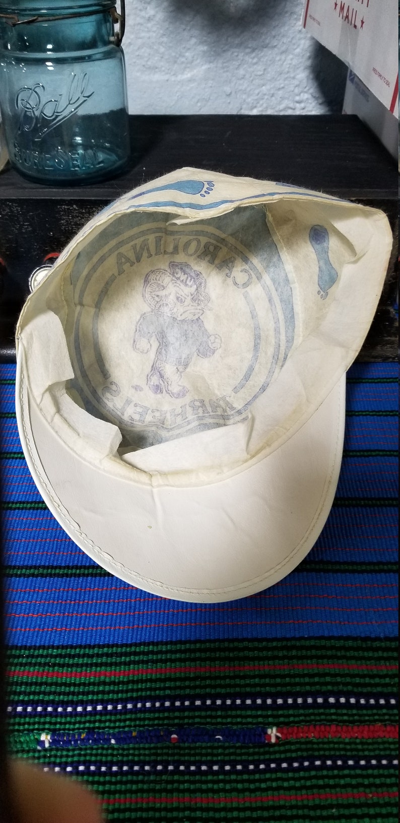 Op de afbeelding: Witte pet met een gebogen klep, met een blauwe afbeelding van een ram en de woorden "CAROLINA TAR HEELS". De binnenkant heeft een bedrukt ontwerp. De pet ligt op een gestreept blauw en groen oppervlak.