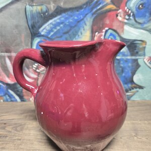May include: A burgundy-colored ceramic pitcher with a curved handle and a spout. The pitcher has a bulbous body and a slightly flared rim. The background features a colorful abstract painting with blue and yellow fish.