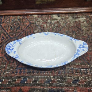 May include: Oval-shaped, white ceramic dish with blue floral patterns around the rim. The dish has two handles and is set against a patterned rug. A wooden cabinet is in the background.