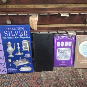 May include: A collection of books on silver, including "Collecting Silver" and "Dealer Guides." The books are in various colors, with the main book featuring silver objects on a blue background. The books are displayed on a wooden surface.