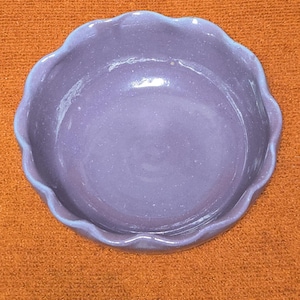 May include: A small, round, lavender-colored ceramic dish with a scalloped edge. The dish is set on an orange surface. The dish is likely used for serving or display.