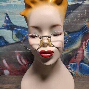 May include: A mannequin head with blonde hair wearing round, silver-rimmed glasses with a clear lens. The mannequin has red lipstick and closed eyes.
