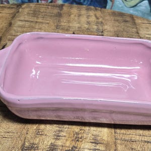 May include: A rectangular, light pink ceramic baking dish with rounded corners and two handles. The dish has a glossy finish and sits on a wooden surface. This item is suitable for baking and serving.