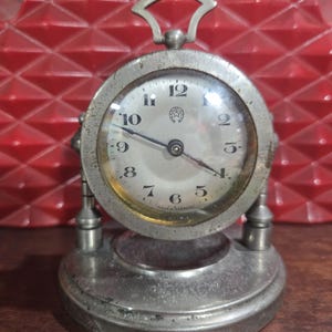May include: An antique silver-toned desk clock with a round face and a handle. The clock face has black numerals and hands, with a small emblem at the top. The clock sits on a circular base with two supporting posts.