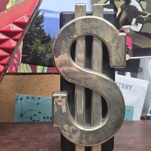 May include: A metallic dollar sign sculpture with a shiny, silver finish. The dollar sign is mounted on a black rectangular base. The sculpture is displayed on a wooden surface, with various posters and artwork in the background.