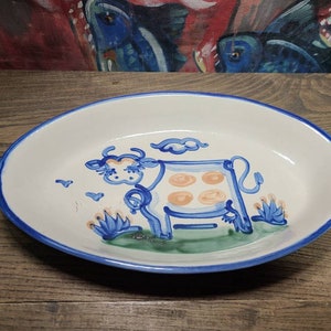 May include: A white oval ceramic baking dish with a blue rim and a hand-painted design of a cow with orange spots standing in a green field. The cow is surrounded by blue and white flowers.