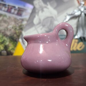 May include: A small, pink ceramic pitcher with a handle. The pitcher has a smooth, glossy finish.