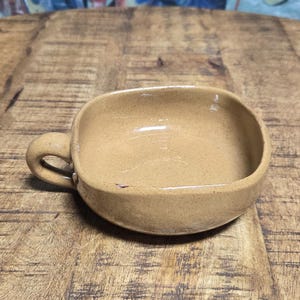 May include: A tan ceramic bowl with a handle. The bowl is square with rounded corners. The background is a wooden surface with a painting of blue fish.