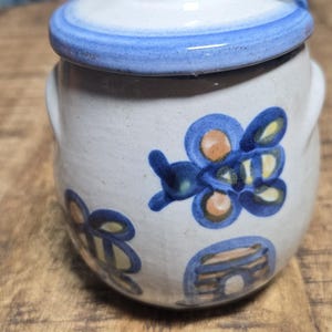 May include: A ceramic jar with a lid, featuring hand-painted blue and orange bee designs. The jar has a white base with blue accents and small handles. The lid is white with a blue rim. The jar is sitting on a wooden surface.