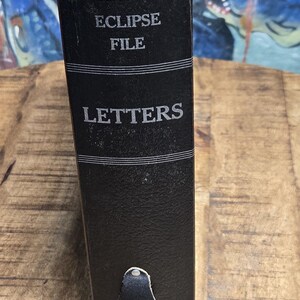 May include: A vintage black Eclipse File Letters binder with silver lettering. The binder has a textured surface and a green top. The Globe-Wernicke Co. logo is visible. A classic office supply.