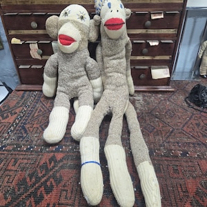 May include: Two handmade sock monkeys. One is wearing a blue and white striped hat. Both have red stitched mouths, tan bodies, and white feet. They are sitting on a patterned rug in front of a wooden cabinet.