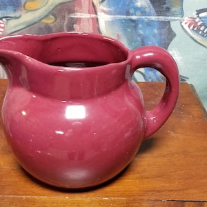 May include: A deep burgundy ceramic pitcher with a rounded body and a slightly flared lip. The pitcher has a simple, elegant design and a smooth, glossy finish.