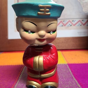 May include: A small ceramic figurine of a person wearing a blue and gold hat and a red robe with gold trim. The figurine has a smiling face with painted on eyes and a single tear coming from each eye.