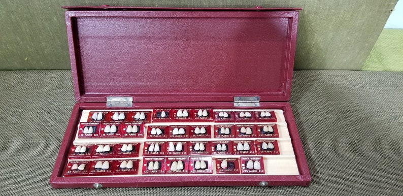 May include: A burgundy case with a hinged lid, open to reveal multiple sets of artificial teeth. Each set is mounted on a small, rectangular card. The case is lined with a cream-colored material.