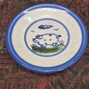 May include: A small, white ceramic plate with a blue rim. The plate features a hand-painted design of a pig with black spots, a blue cloud, and green grass.