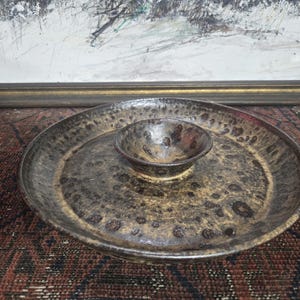 May include: A ceramic serving dish with a central dip bowl. The dish is a mottled brown and tan color with a speckled pattern. The bowl is a smaller version of the same design. The dish is sitting on a patterned surface.
