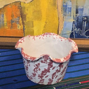 May include: A white ceramic bowl with a scalloped rim, decorated with maroon splatters. The rim is painted maroon. The bowl sits on a blue and green striped textile, with a painting in the background.