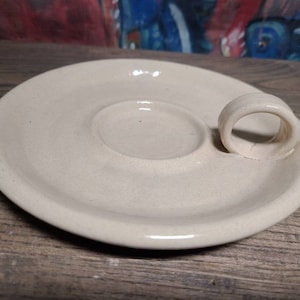 May include: Off-white ceramic plate with a circular indentation in the center and a curved handle. The plate has a smooth, glossy finish and sits on a wooden surface. The design is simple and functional, suitable for serving small portions.