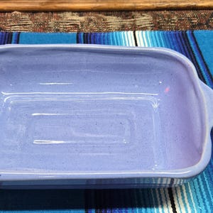 May include: A rectangular, light purple ceramic baking dish with rounded corners and two handles. The dish is empty and sits on a blue and white striped textile. The baking dish is a kitchenware item.