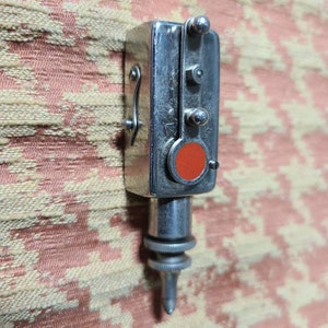 May include: A silver-colored metal tool with a red button and a pointed tip. The tool has a small lever on the side and three small knobs on the front.