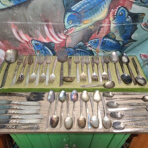 May include: A collection of vintage silverware, including forks, spoons, and knives, arranged on a green surface. The silverware is made of silver and has a polished finish.
