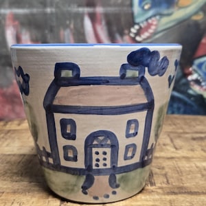 May include: A ceramic planter featuring a hand-painted blue and brown house design on a cream background. The vessel has a tapered shape with a blue rim, depicting a house with a chimney, windows, a door, and a path, set against a light green base.