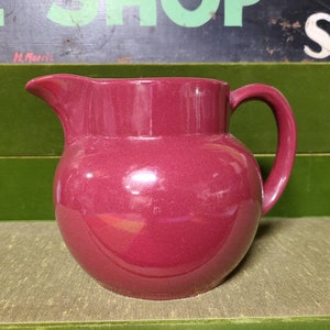 May include: A deep burgundy ceramic pitcher with a rounded body and a handle. The pitcher is glazed and has a smooth, shiny finish.