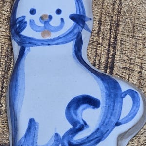 May include: A white ceramic cat ornament with hand-painted blue outlines and details. The cat features pointed ears with tan accents, a simple facial expression with blue whiskers, and a curled tail. A small hole is located at the top for hanging.