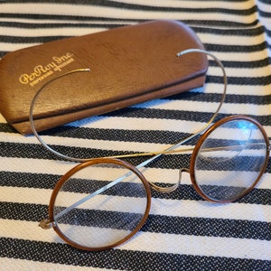May include: A pair of round, brown-rimmed glasses with a silver metal frame and nose bridge. The glasses are resting on a striped fabric surface. The glasses are in a case that says "PerRoy Inc. dispensing opticians".