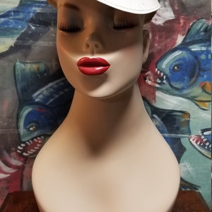 Op de afbeelding: Een mannequinhoofd met een wit en blauwe pet met het woord "HEELS" erop gedrukt. De pet heeft een witte rand. De mannequin heeft gesloten ogen en rode lippenstift.