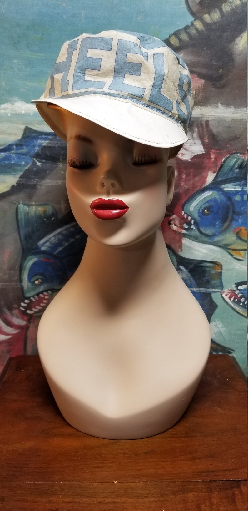 Op de afbeelding: Een mannequinhoofd met een wit en blauwe pet met het woord "HEELS" erop gedrukt. De pet heeft een witte rand. De mannequin heeft gesloten ogen en rode lippenstift.