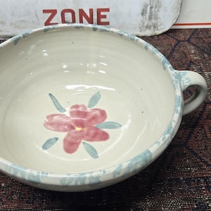May include: A cream-colored ceramic mixing bowl with a single handle and a pouring spout. The interior features a hand-painted pink flower with green leaves at the center, while the rim is decorated with a light blue sponge-painted pattern.