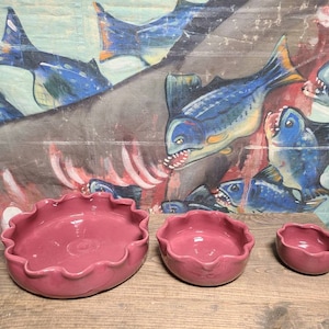 May include: Three pink ceramic bowls with scalloped edges. The bowls are arranged in a row from largest to smallest on a wooden surface.