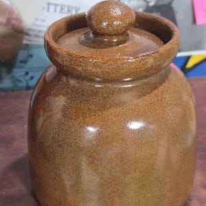 May include: A brown ceramic jar with a lid. The jar has a rounded body and a slightly textured surface. The lid is a matching brown color and has a small, round knob on top. The jar appears to be handmade and has a rustic aesthetic.