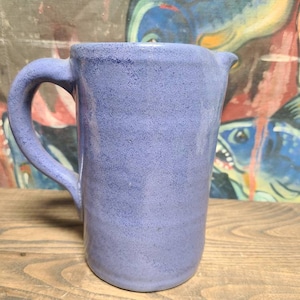 May include: A light blue ceramic pitcher with a textured surface and a handle. The pitcher is sitting on a wooden surface.