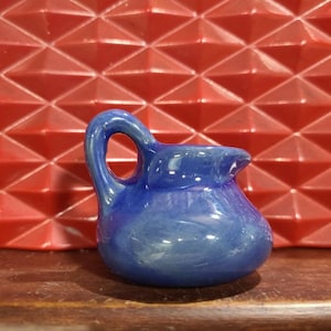 May include: A small, blue ceramic pitcher with a handle. The pitcher is glazed and has a shiny finish.