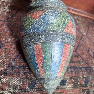 May include: A small, ceramic vase with a pointed bottom. The vase is decorated with a geometric pattern of red, blue, and green stripes. The vase has a speckled, textured surface.