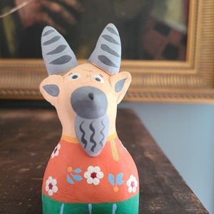 May include: A hand-painted ceramic goat figurine with a gray beard, orange shirt with white flowers, and green pants. The goat has gray horns and large, white eyes.