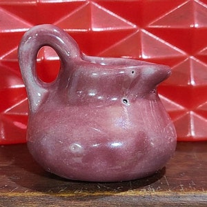 May include: A small, purple ceramic pitcher with a handle. The pitcher has a smooth, glossy finish and a slightly rounded shape.